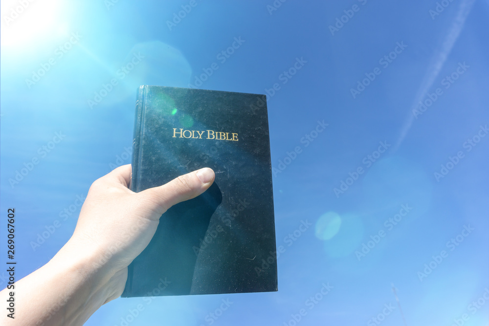 Wall mural hand holding holy bible against a blue sunny sky