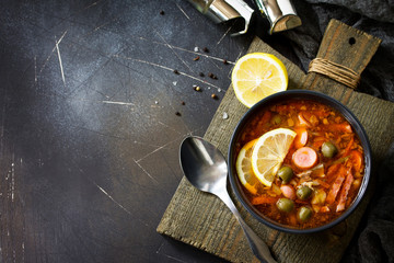 Traditional Russian soup Solyanka with meat, sausages, vegetables, capers, pickles and olives with lemon on black background. Rustic style. Top view flat lay background. Free space for your text.