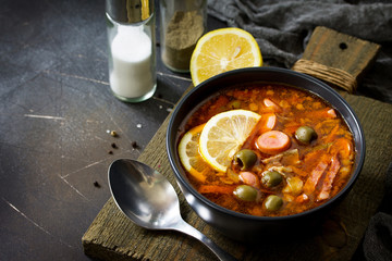 Traditional Russian soup Solyanka with meat, sausages, vegetables, capers, pickles and olives with lemon on black background. Rustic style. Free space for your text.