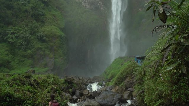 Woman walking across a bridge at the base of Sipiso Piso waterfall with a tilt reveal the beautiful waterfall in the background.