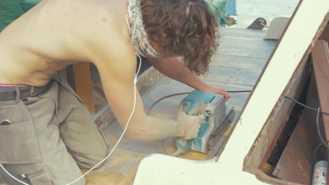 Man sands teak deck planking with belt sander on wooden boat topless in Summer sunshine.