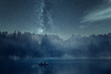Nachtangeln auf einem See bei Nacht mit Sternenhimmel und Nebel. Im Hintergrund Wald, Berge und die...