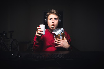 Young man sitting at the table in the room eats chips and holds drink in his hands, looks at the computer screen,astonished face.Surprised teen eats snack, watches a scary video on a computer at night