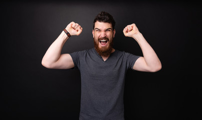 Bearded man over dark background celebrating and screaming with rised hands