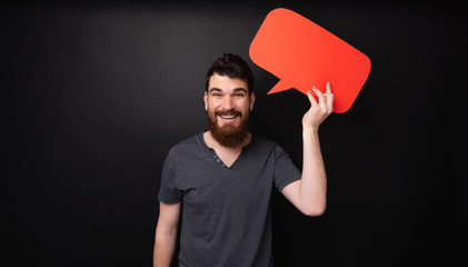 Cheerful bearded man holding red  empty bubble speech  over dark isolated background