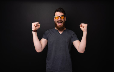 Naklejka premium Photo of bearded man wearing sunglasses celebrating with rised hands standing over dark background