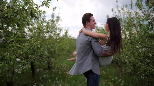 Closeup of groom circling bride carrying in arms, joyful barefoot woman kissing beloved man hugging by neck. Attractive mixed race couple enjoying engagement date in apple garden among flowering trees