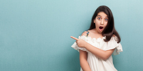 Portrait of beautiful brunette young girl with long straight hair in white dress standing, pointing at wall empty copyspace looking at camera with shocked face. studio shot isolated on blue background