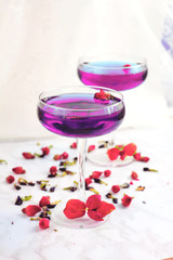 Thai asian purple Anchan tea in cocktail glasses with dry rosebuds and blue tea, tea leaves and green basil leaves, healthy organic detox Asian herbal tea clitoria Butterfly Pea on marble background