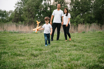 Fototapeta premium Father, mother and son playing with toy airplane in the park. friendly family. People having fun outdoors. Picture made on the background of the park and blue sky. concept of a happy family.