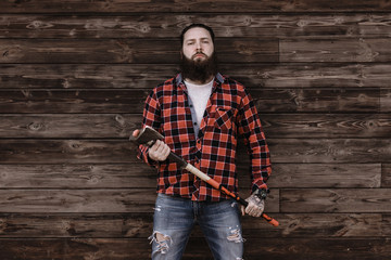 Brutal strong man with a beard dressed in a checked shirt and torn jeans stands with an ax in the hands on the background of a wooden wall