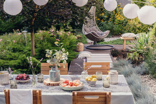 Lanterns above table with flowers, food and drink in the garden with hanging chair and plants. Real photo