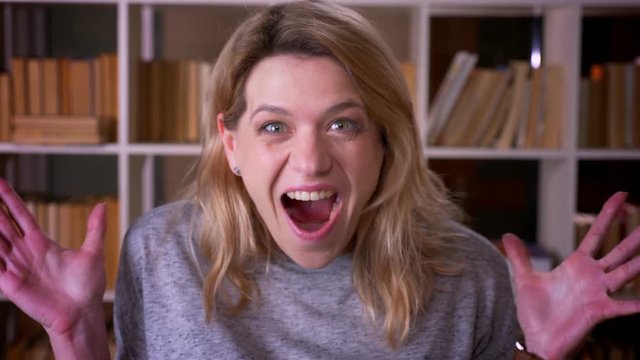 Middle-aged blonde teacher shows strong amusement and happiness into camera coves face with hands at the library.