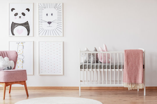 Pink chair near cradle with blanket in white baby's room interior with posters and rug. Real photo