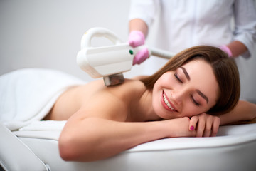 Young pretty smiling woman back laser epilation in beauty salon. Cosmetologist doing depilation treatment with elos device. Beautician hands in gloves holding hair removal cosmetology ipl hardware