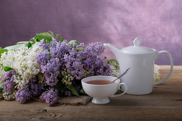 Bouquet of lilac, a cup of tea, a white teapot on a wooden table.
