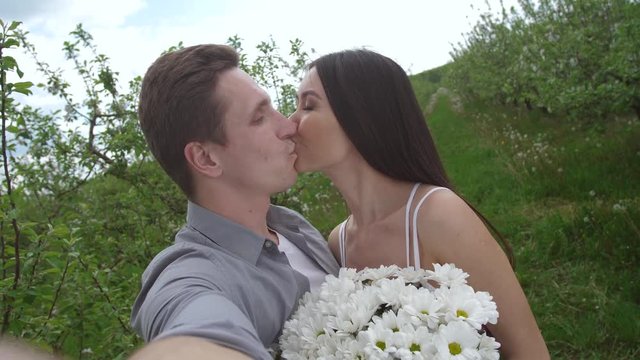 Close-up of joyful mixed race couple taking selfie photo of their romantic kiss on background of flowering trees in spring orchard. Loving bride and groom kissing enjoying togetherness and closeness
