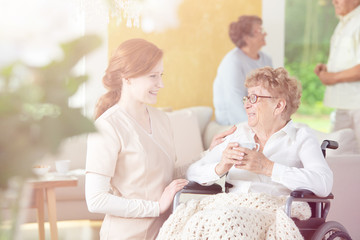 Friendly caregiver helping smiling senior woman in the wheelchair in the nursing house