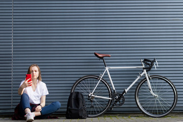 beautiful young girl with a smartphone, a backpack and a bicycle sits on the background of the wall, she stopped to rest