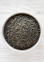 Dry green french lentils in a bowl over white wooden surface, top view. Flat lay, overhead, from above. Close-up.