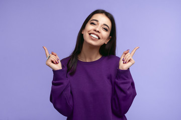 Pretty adult girl with broad smile, wears purple sweatshirt, pointing fingers up at copyspace,...