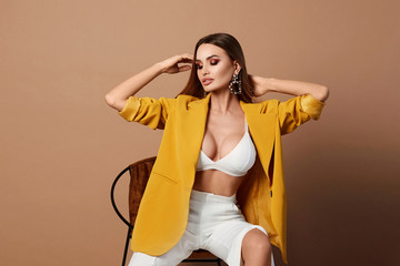 Beautiful and fashionable blonde girl with the sexy perfect body, with big bust and with full lips, in the stylish yellow blazer posing at the orange background