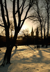 Two people walk in a park at sunset in Tallinn, Estonia with St Charles' Church in the background and the trees casting long shadows on the snow