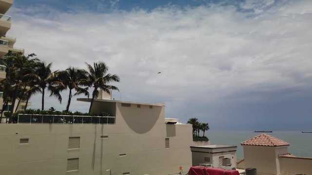 Pan of the sky following a military fighter jet aircraft seen flying fast over the ocean near a tropical beach and then past a hotel. Palm trees are seen.     