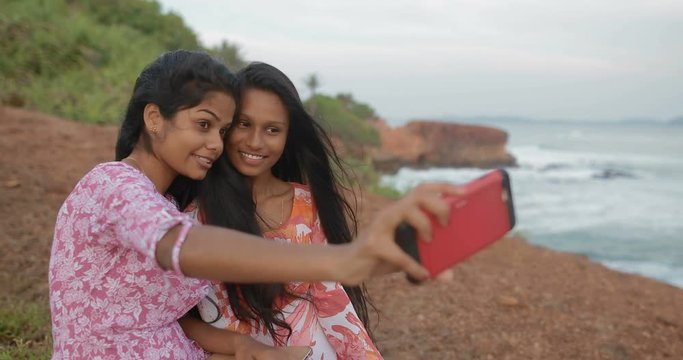 Two Young South Asian attractive girls sitting near the ocean using smartphone. Girlfriends take pictures on the smartphone. real time. Shot on Canon 1DX mark2 4K camera