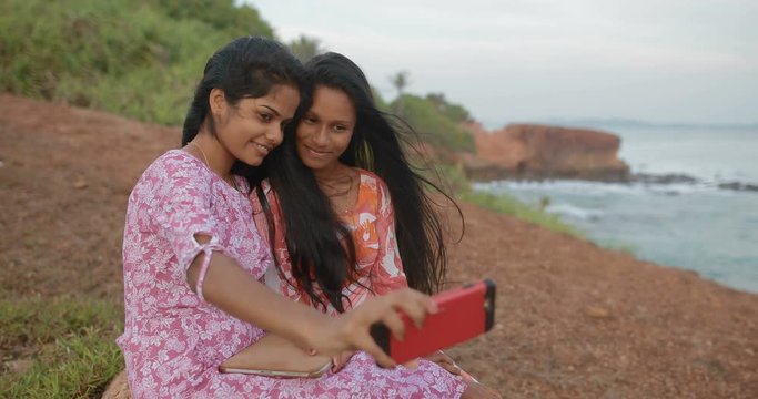 Two Young South Asian attractive girls sitting near the ocean using smartphone. Girlfriends take pictures on the smartphone. in slow motion. Shot on Canon 1DX mark2 4K camera