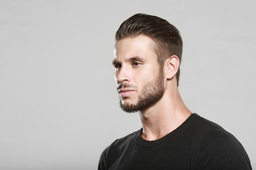 Obraz premium Muscular model young man with beard in black t-shirt on gray background. Fashion portrait of brutal sporty sexy strong muscle guy with modern trendy hairstyle. Model, fashion concept.