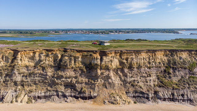 An aerial view of a coastguard tower on the top of a hill with eroded cliff along a sandy beach with an harbor in the background under a majestic blue sky and some white clouds