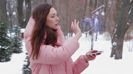 Beautiful young woman in a winter park interacts with HUD hologram gift box. Red-haired girl in warm pink clothes uses the technology of the future mobile screen - Powered by Adobe