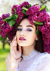 Portrait of a young beautiful girl in a wreath of lilacs. White lace dress on the perfect figure of the model. Photo session in the spring garden in the open.