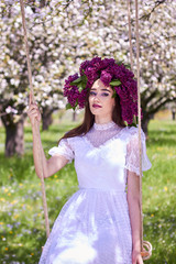 Obraz na płótnie Canvas Young beautiful girl in a wreath of lilac, sits on a makeshift swing. White lace dress on the perfect figure of the model. Photo session in the spring apple orchard in the open.