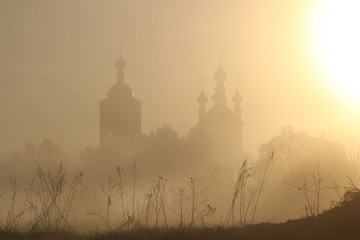 Fototapeta na wymiar Misty rural landscape in rising sun light with silhouettes of church and grass 