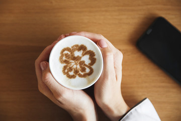 Glass with art cappuccino in female hands. The phone is near