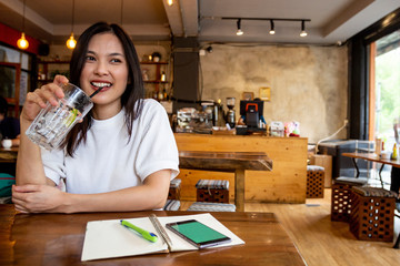 Fototapeta premium Charming beautiful pretty smile Asian woman mobile phone and take note during rest in coffee shop, happy Asian female girl drinks lemonade and use cell telephone while relaxing in cafe free time