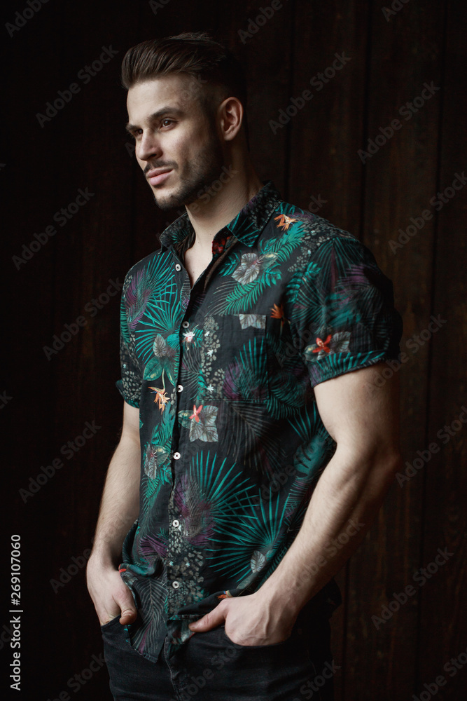 Wall mural Muscular model young man with beard in tropical print shirt on dark background. Fashion portrait of brutal sporty sexy strong muscle guy with modern trendy hairstyle. Model, fashion concept.