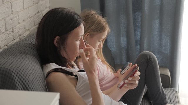 Young mother and daughter are using gadgets sitting on the couch in the living room. Concept modern family during weekend at home. Contemporary technology.