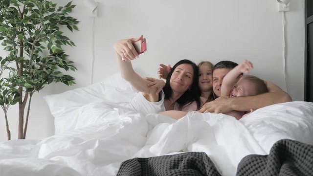 Cheerful young family making funny selfie while laying in bed with mobile phones.