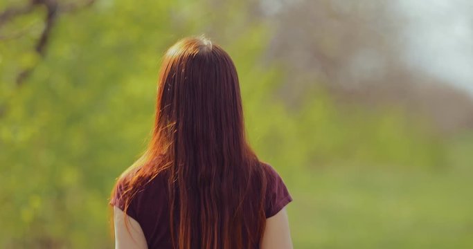 Brunette girl with long hair walks through the park on a sunny spring day. Back view.