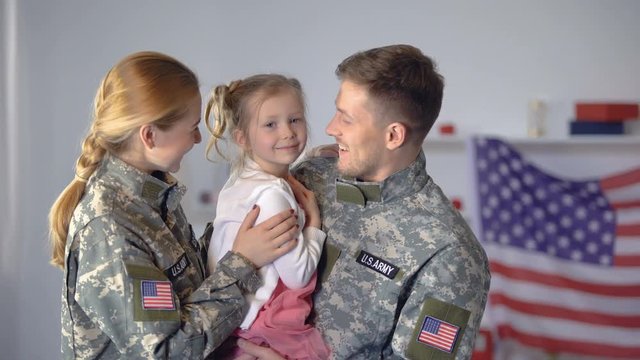 Cheerful military couple kissing little child in cheeks, USA flag on background