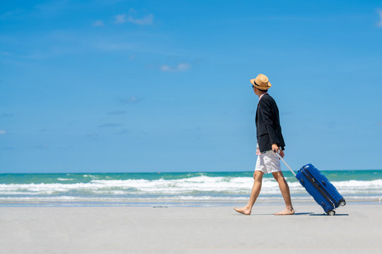 Selective focus of young businessman traveler in casual wear walking with holding blue suitcase on the island beach in summer day holidays vacation with blurred blue sea, sky and clouds backgrounds.