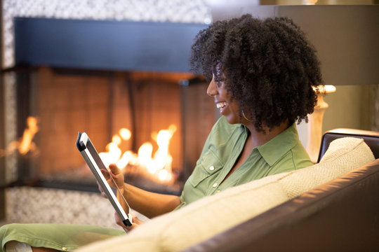 Black African American female relaxing at home and reading or browsing the internet with a digital tablet in a living room by the fireplace.  The image depicts technology in domestic lifestyle.