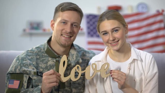 Smiling american soldier and his girlfriend showing wooden love sign on camera