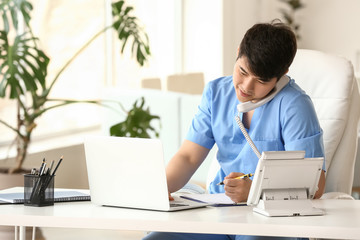 Male medical assistant talking by phone while working on laptop in clinic