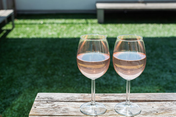 Two glasses of rose wine on a wooden table in sunlight top view with a garden bench in the background