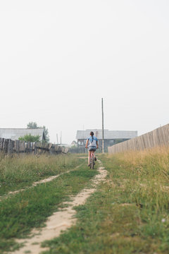white caucasian young woman in casual clothing riding on bicycle in countryside at morning sunrise, view from back, vertical stock photo image
