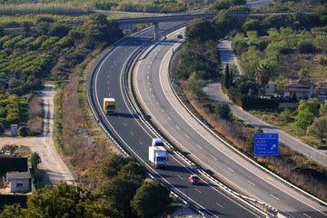 Fototapeta na wymiar A long curve of the AP7 motorway in Spain near Denia, photographed from above with cars and trucks. On the right is a blue sign for the next departure.